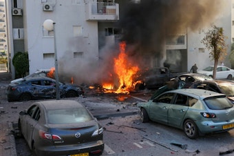 caption: A member of the Israeli security forces stands near burning cars following a rocket attack from the Gaza Strip in Ashkelon, southern Israel, on Oct. 7.