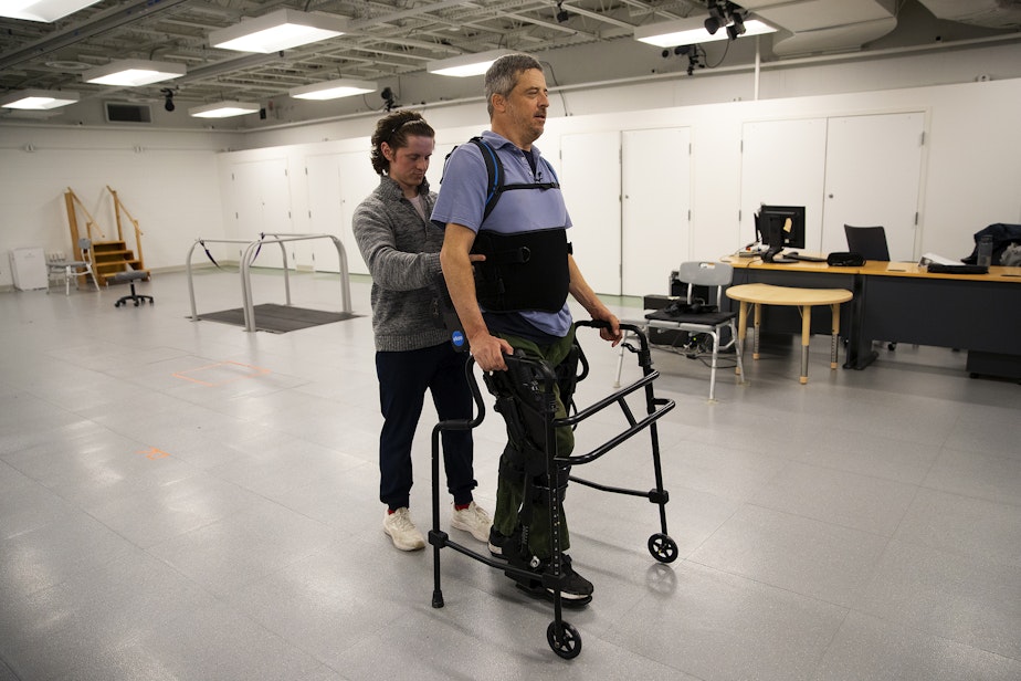 caption: Jon Schlueter, who became injured in a diving accident, walks in an eksobionics device with the help of Rich Henderson, a graduate student and physical therapist, on Monday, April 15, 2024, at Wallace Hall on the University of Washington campus in Seattle.