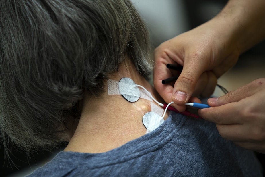 caption: Research assistant Adrià Robert Gonzalez places electrodes on the back of Debbie Strom's neck before a non-invasive transcutaneous spinal cord simulation session on Monday, April 15, 2024, at Wallace Hall on the University of Washington campus in Seattle.