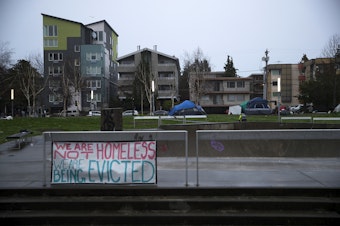 caption: 'We are not homeless, we are being evicted,' reads a sign in the foreground as Seattle Parks and Recreation employees prepare to sweep unhoused community members from Ballard Commons Park on Tuesday, December 7, 2021, in Seattle. 